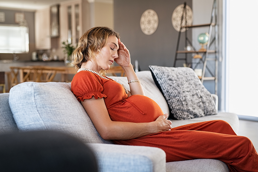 Exhausted middle aged pregnant woman suffering headache sitting on couch in the living room. Stressed young pregnant woman with terrible migraine. Expecting mother holding belly bump feeling worrying.